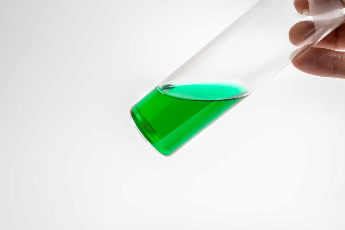 緑色の薬液が入った試験管を手に持つ様子。化学物質管理やリスクアセスメントに関連する現場での確認作業の一場面を表現。