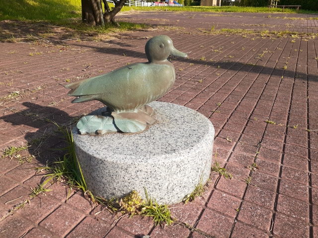 公園のレンガ舗装上に設置されたカモの銅像。円形の御影石台座の上に載っており、足元にはハスの葉の装飾がある。